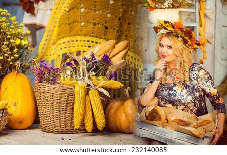 girl holding fresh pastries. Yellow maple wreath on his head. Basket of  flowers and bread, big pumpkins in room with autumn interior.