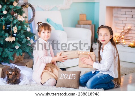 two teenage girls are sitting by the Christmas tree and the fireplace and give each other gifts