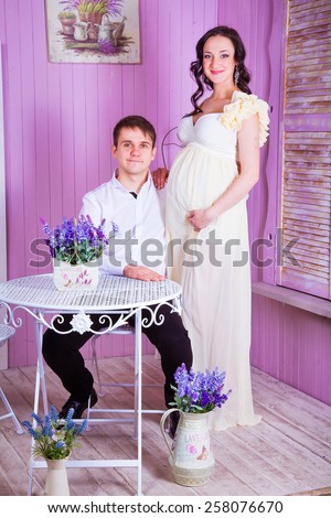 pregnant wife in a long light dress with her husband  sitting at a table in a pink interior Studio