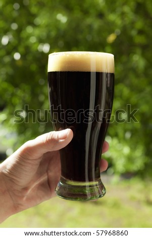 Man\'s hand holding glass of dark foam beer on background of green summer foliage