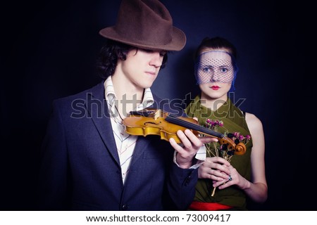 young man in hat with violin and young woman in veil with bouquet of roses on black background