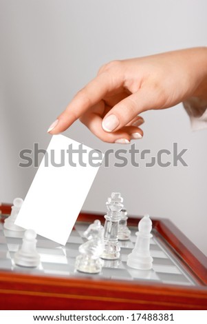 Woman hand holding blank business card  (visit card) over a chessboard with glass figures. Closeup.