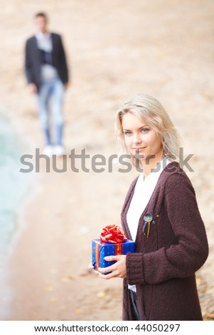 Young couple having fun at the beach. Girl holding a gift. Boyfriend walking on a background. Focus on a girl.