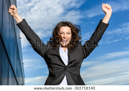 Young Business Woman is jumping for joy in front of blue cloudy sky with office block to the side