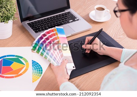 image of a graphic designer using graphic tablet in her work on the foreground and working with color samples for selection. A concept of modern digital workplace