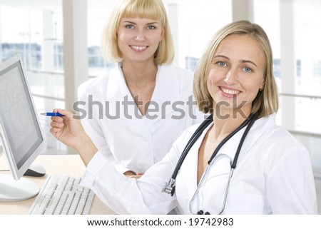 Two female physicians  stand near a computer and one of them points at a monitor.