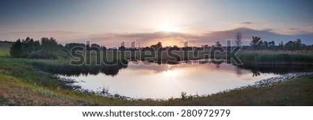 Summer rural landscape with river at dawn. Village on banks of river and beautiful rays of sun in sky. Panorama. Mirror reflection in water