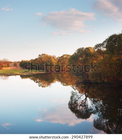 Spring landscape with river. Trees on banks of river reflected in water. Sunny evening. Reflection of cloudy sky in water