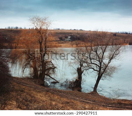 Rural landscape with frozen river. Lonely house on hill on cloudy day. Early spring. Sad mood