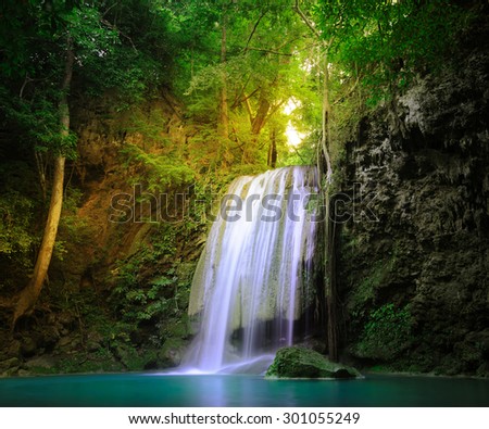 Amazing nature place. Sunlight beams and rays shining through wild jungle forest trees and plant leaves around tranquil waterfall falling in natural pond