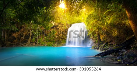 Exotic background of beautiful jungle forest with majestic waterfall falling into blue water lake and sun light rays shining through green leaves of tropical plants ant trees. Rainforest panorama