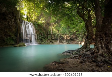 Sunlight beams and rays shine through leaves of trees in tropical rainforest park in Thailand with beautiful waterfall falling in clear pond and old big tree on foreground