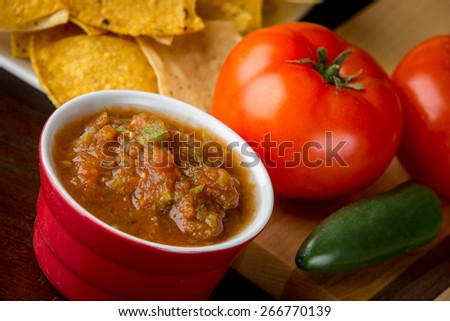 Fresh tomato salsa surrounded by ripe tomatoes and jalapeno. In the background is corn chips.
