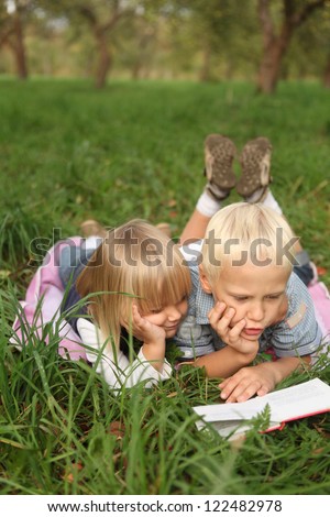blond 6-year-old boy and 4-year-old girl reading a book lying on the grass.
