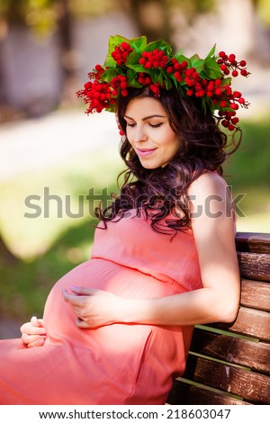 Portrait of pregnant women in the dress with wreath in her head  sits on bench in the warm park