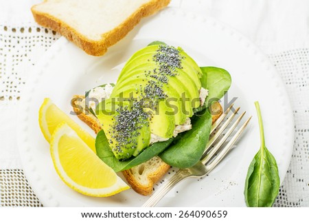 Sandwich of the pair of slices of fresh white bread toast with spinach, chicken, dressing, sliced avocado and poppy seeds. On a white plate and white vintage embroidered tablecloth. selective Focus