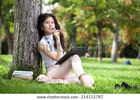 Young woman caucasian asian stand leaning against a tree in the park and thinking what to write in her agenda