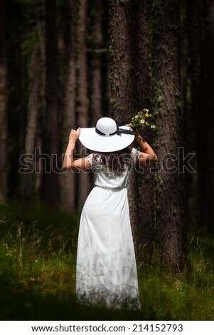 Woman from behind in white dress and white hat walking in the wood holding a bunch of flowers