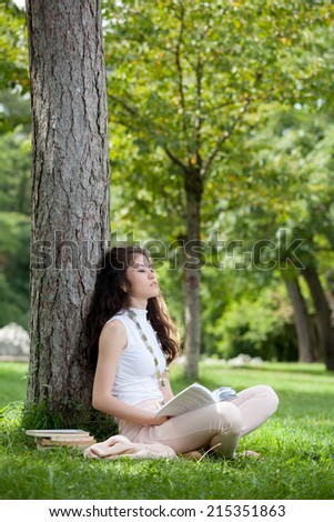 Young woman caucasian asian dreaming laying against a tree sitting in the grass with a book in hands
