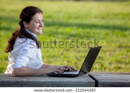 Smiling business mature woman writing on laptop sitting on the wooden table in the park