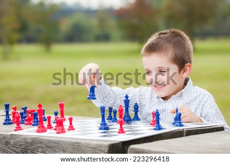 Elegant young boy in white shirt learning to play chess with blue and red chess pieces on wood table in the park