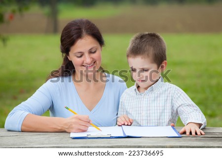 Young woman teacher teaches little young boy in white shirt writing or drawing with a pencil on a sheet of paper on wood table in the park
