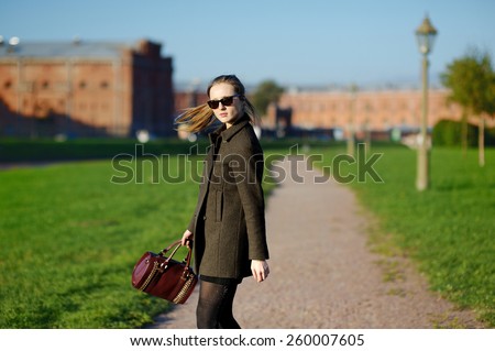 Lovely young woman, blond, slender, with long straight hair, glasses, coat, posing in the park on a background of alleys and architectural structures