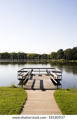 Color photograph of an empty dock with benches on a small lake.