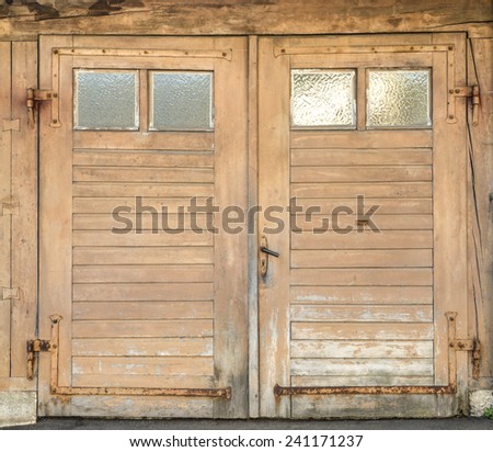 Old, light brown, dirty and weathered wooden garage door with four small glass windows and rusted iron fittings.
