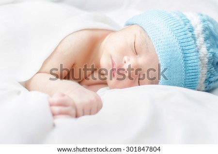 Sleeping New born baby boy wearing blue hat covering silk white bed sheet
