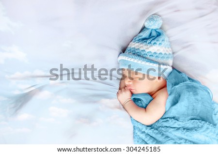 Newborn baby boy sleeping on white silk bed floating clouds wearing blue hat and blanket isolated in white background