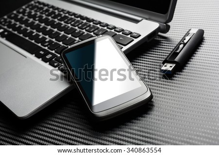 Blank Black Smartphone With Blue Reflection Leaning On A Notebook Keyboard Next To A Plugged In USB Storage Flash Drive, All Above A Carbon Layer