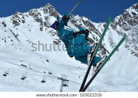 a ski jumper performing a 360 degree spin in the air, and preparing to land. In the background ski lifts, ski runs and snow covered mountains