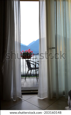 a gentle breeze wafts the curtains of a hotel room with balcony. through the open windows a romantic view of a lake in the mountains