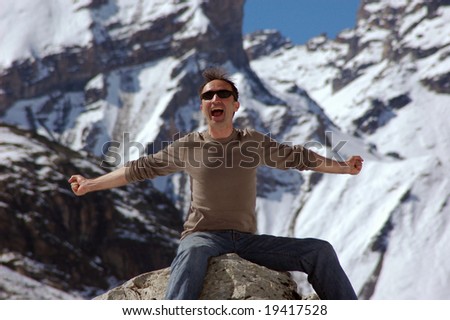 Happy man perched on rock in the swiss alps with arms stretched out; Photo shot in the afternoon;  In the background snow and glaciers of the swiss alps;