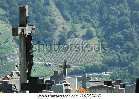 Statue of Jesus on a cross in a graveyard in a swiss mountain village. Around the foot of the statue, a cluster of gravestones, and in the background vineyards, producing the blood of christ.