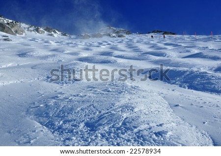 skiers crossing the barrier onto an off-piste mogul field on a very steep glacier. Plumes of blown snow are lifted from the summit