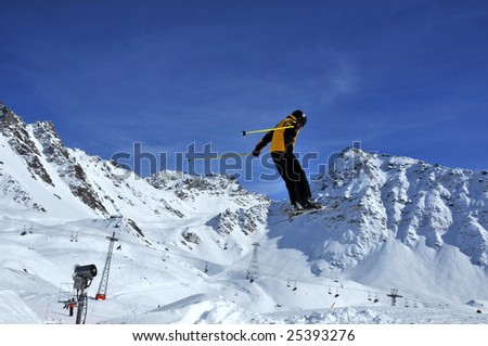 aeroski: a skier in yellow and black performing a high jump against a backdrop of mountains and ski lifts