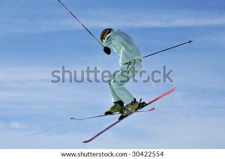 Aeroski:  a skier with pale green clothes performing a tele-heli  (crossed skis in the air) in the sky