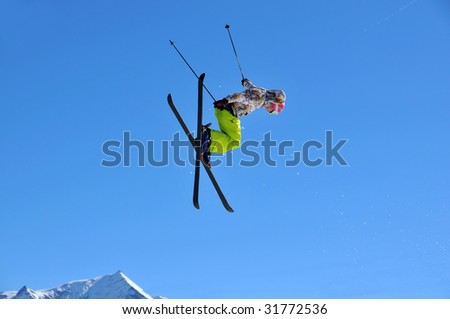 a girl skier performing a high jump with her skis crossed (tele-heli). In the background snow covered mountains