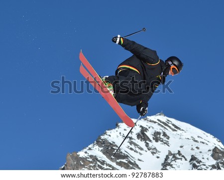 a young free rider on skis performing a high jump. In black clothes and with pink skis, trailing snow. Against a clear blue sky