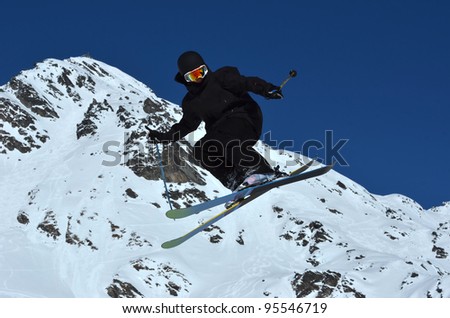 a free ride skier dressed all in black, makes a high jump against a backdrop of a clear blue sky and snow covered mountain