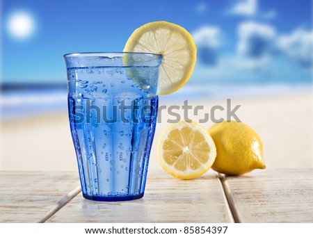 a Blue glass with sparkling water and lemon on a wooden deck overlooking a tropical beach - focus on the lemon