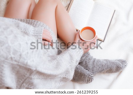 Soft photo of a woman in a cozy sweater on the bed with the old book and a cup of tea with milk in their hands
