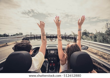 Friends having fun at car trip around the world. Couple in love with arms up on a convertible car.
