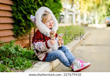 Little girl with a hood on her head eating ice cream while sitting on the sidewalk. Her mouth smeared with ice cream