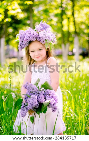Little beautiful blonde girl wearing a white dress with a bouquet of flowers in her hands and a diadem of lilac flowers on the head stands in the park on a sunny morning and dreams.