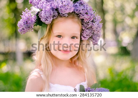 Little beautiful blonde girl wearing a white dress with a bouquet of flowers in her hands and a diadem of lilac flowers on the head stands in the park on a sunny morning and smiling.