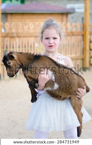 Little beautiful blonde girl standing on a farm yard, holding a brown goat in her arms and smiling mischievously.