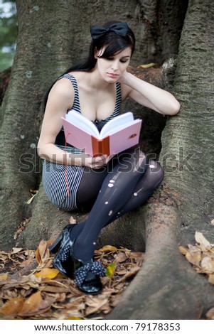 Portrait of a beautiful young woman with long brunette hair sitting under a huge tree with a pink notebook